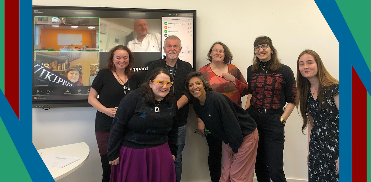 A photo of a smiling group of people in front of a screen showing people who joined the training remotely.