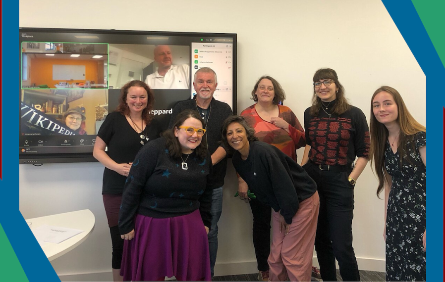 A photo of a smiling group of people in front of a screen showing people who joined the training remotely.