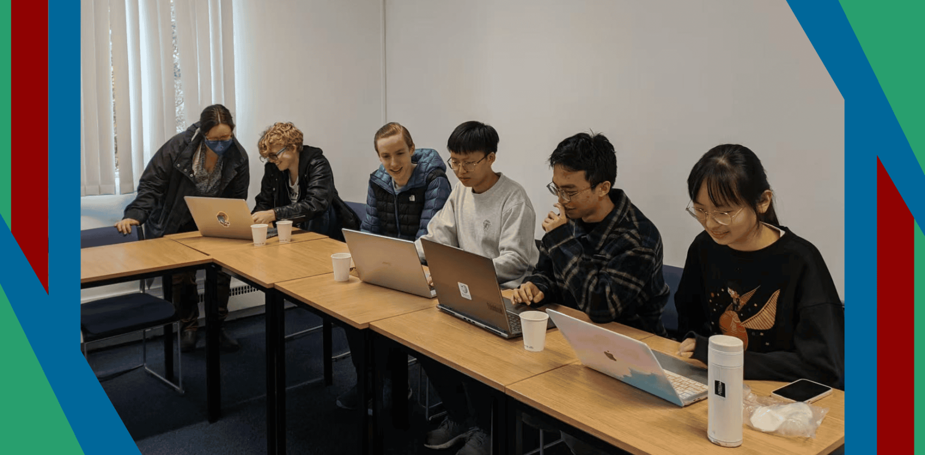 A photograph of a group of university students sat at a desk working on their laptops, framed by the Wikimedia UK colours of green, red and blue.