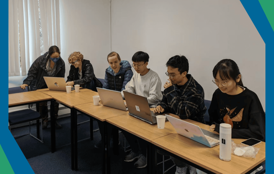 A photograph of a group of university students sat at a desk working on their laptops, framed by the Wikimedia UK colours of green, red and blue.
