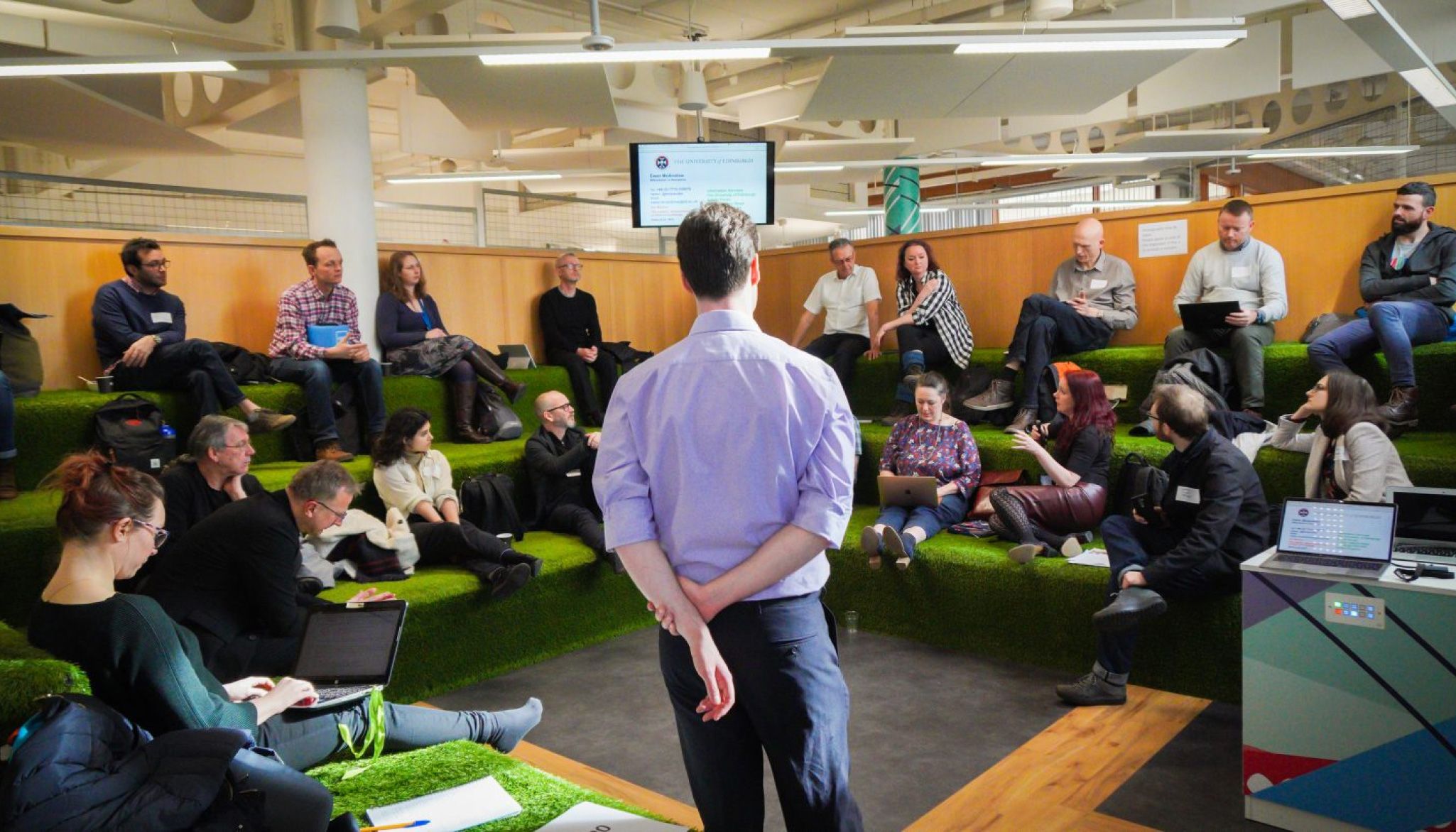 A speaker addresses a group sitting on tiered, grass-covered seating at a workshop