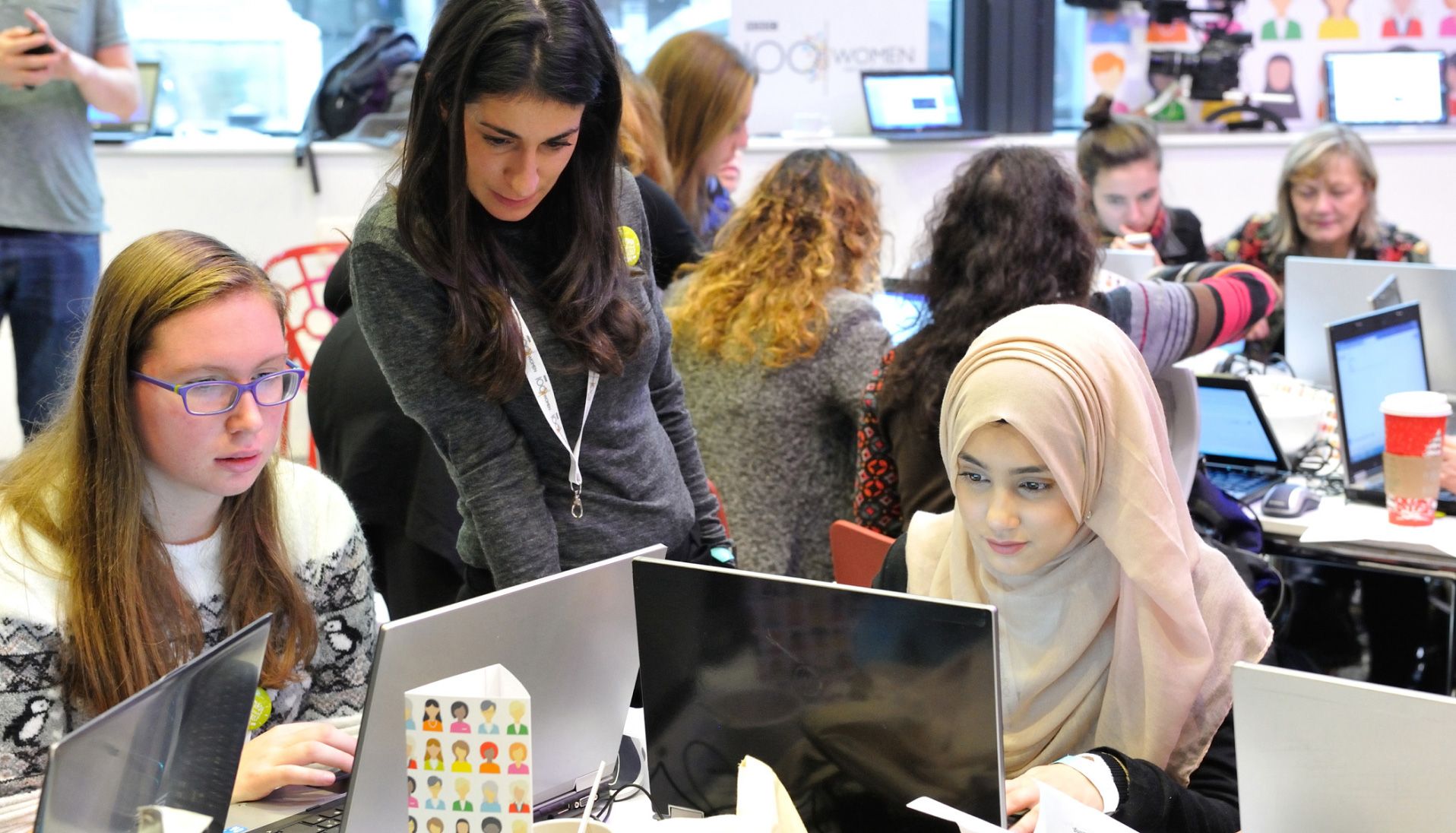 Women working on laptops at a Wikipedia editathon, with a volunteer leaning over to help a participant